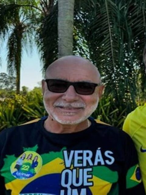 Fabrício Queiroz sorrindo ao ar livre, de óculos escuros e camiseta com a bandeira do Brasil, entre palmeiras